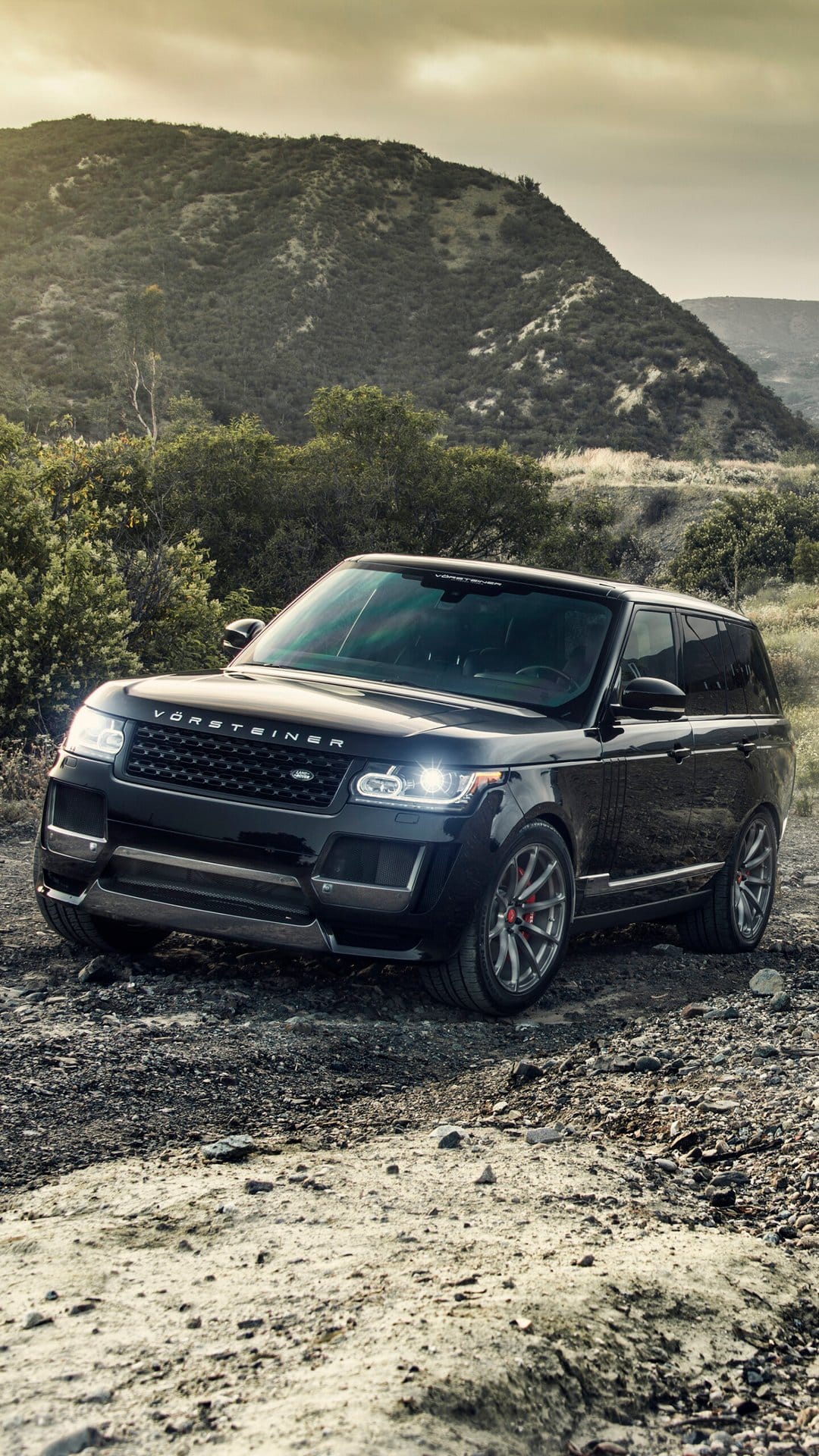 Black Vorsteiner SUV with headlights on parked on a rocky path against a large hillside under a cloudy sunset.