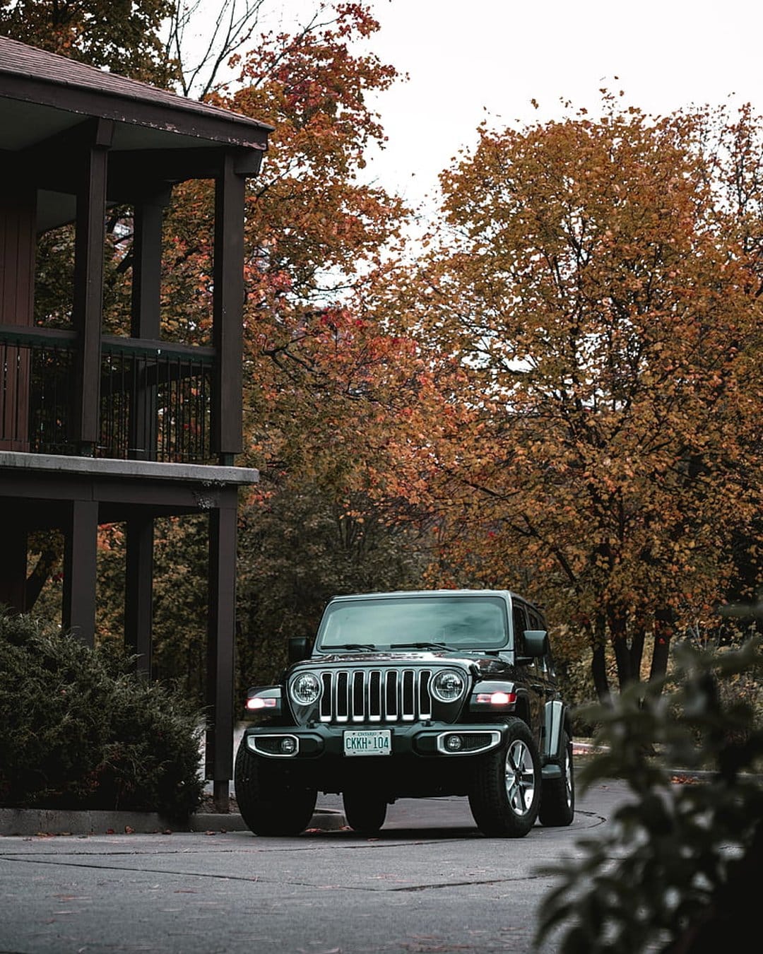 Dark green Jeep Wrangler parked on a paved driveway surrounded by orange autumn trees and a rustic building.