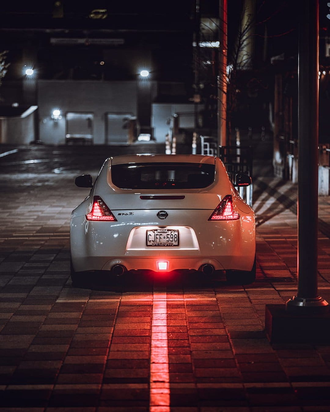White Nissan 370Z rear view with glowing red brake lights parked on a brick street under urban night lighting.