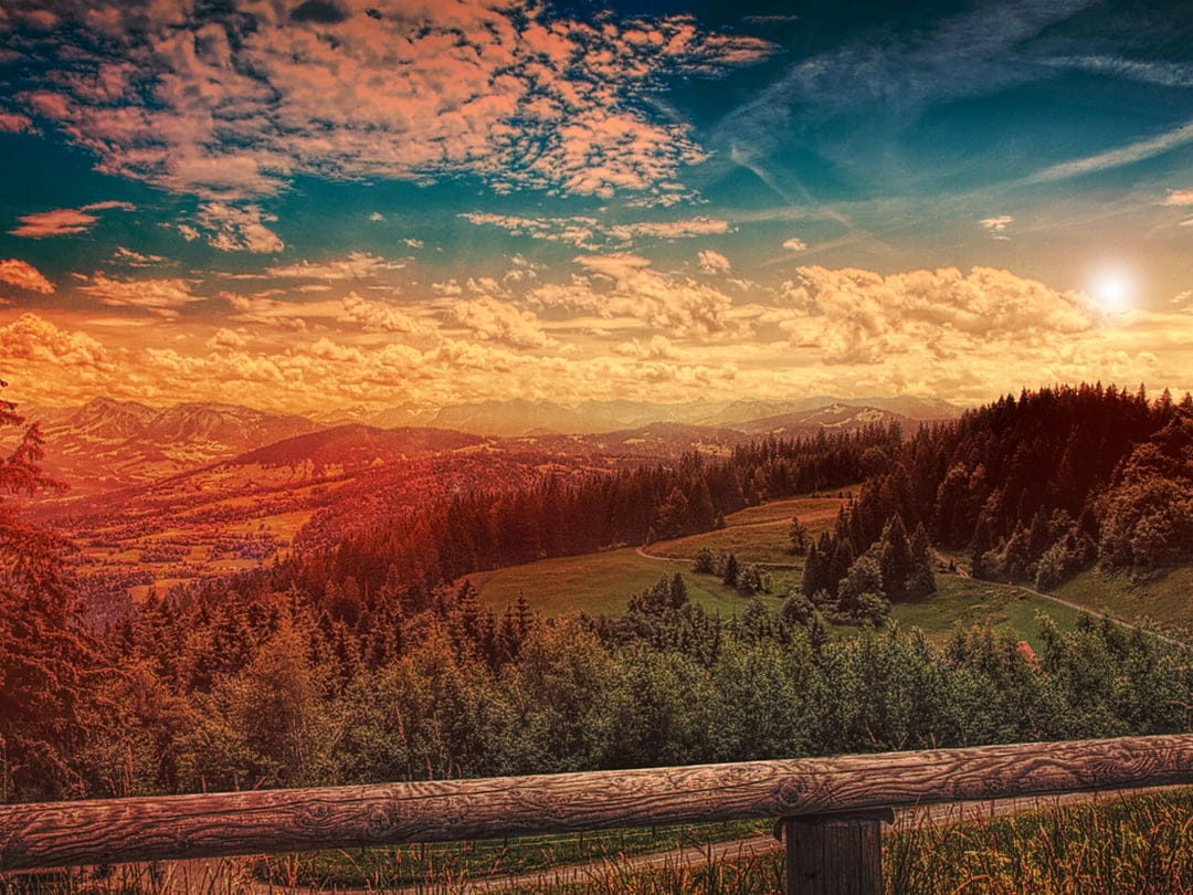 Rolling mountains under an orange sunset sky with a rustic wooden fence framing the foreground landscape.