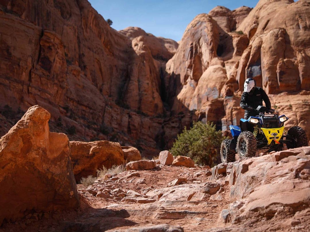 Blue and yellow ATV with a helmeted rider on rocky sandstone terrain against massive red canyon walls.
