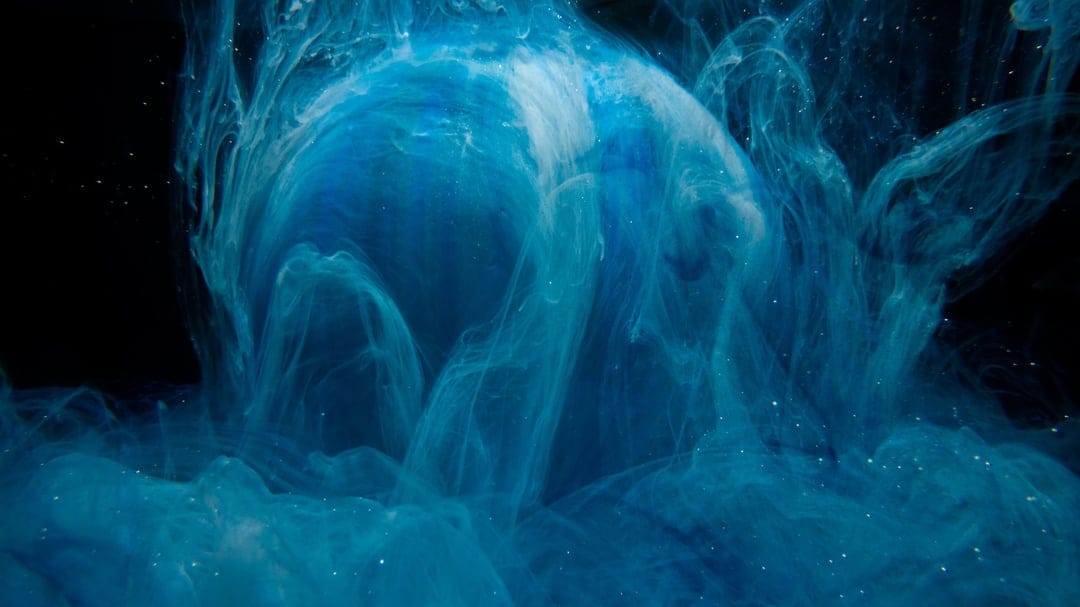 Glowing white sphere surrounded by turbulent swirls of cobalt blue ink and fine particles on a dark backdrop.