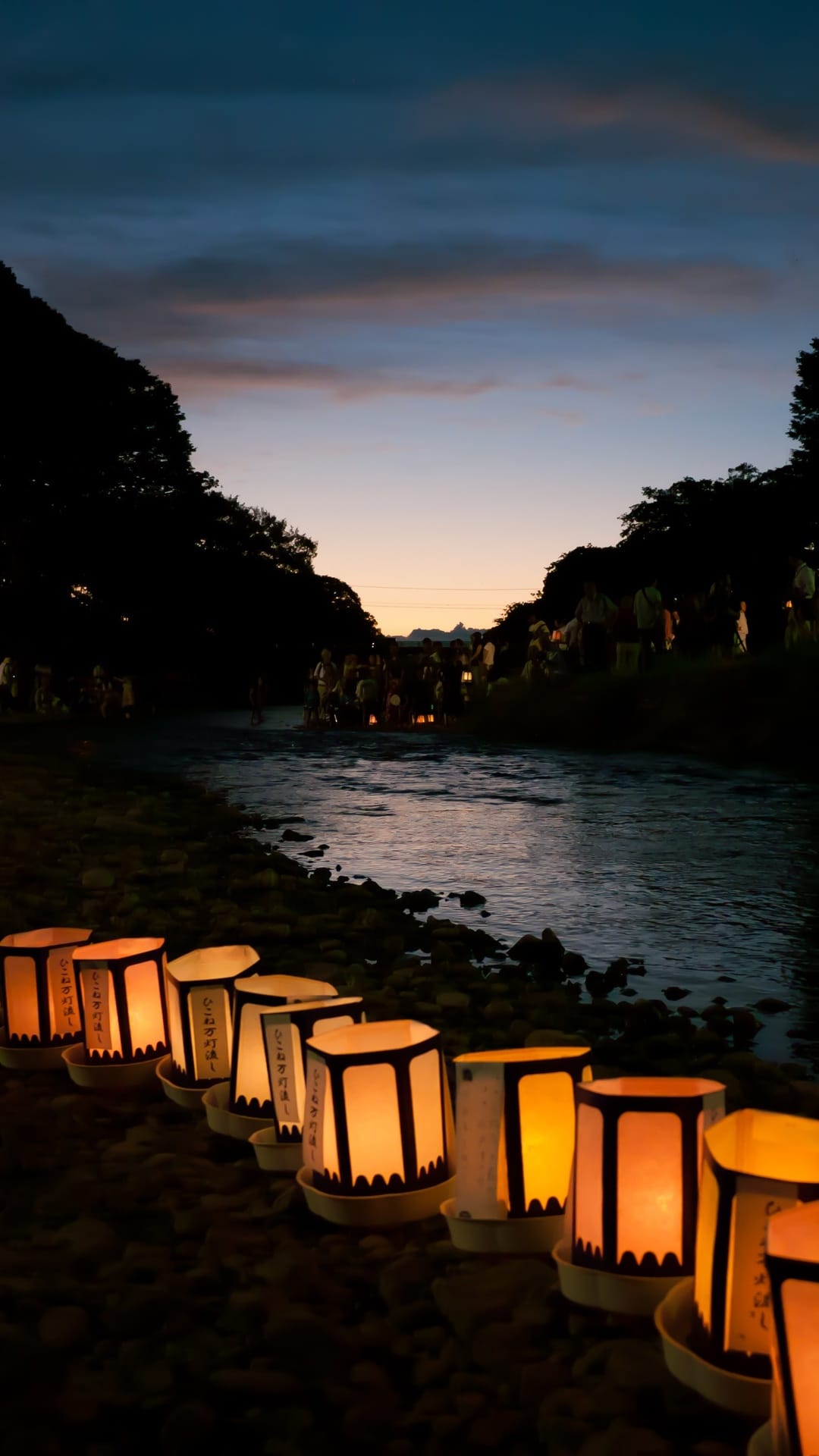 Paper lanterns glow on a riverbank with reflections on the water surface and silhouetted trees in the background.