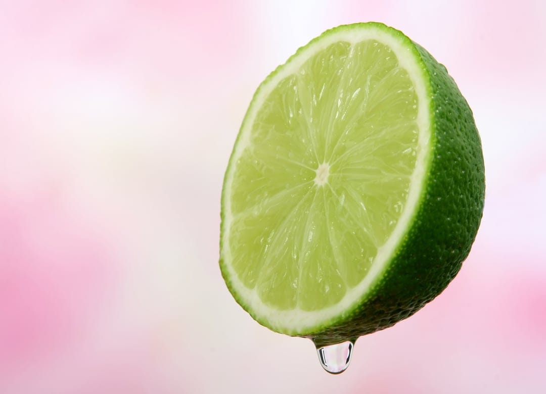 Lime slice dripping a single water droplet against a blurred pink and white background in a vertical macro shot.