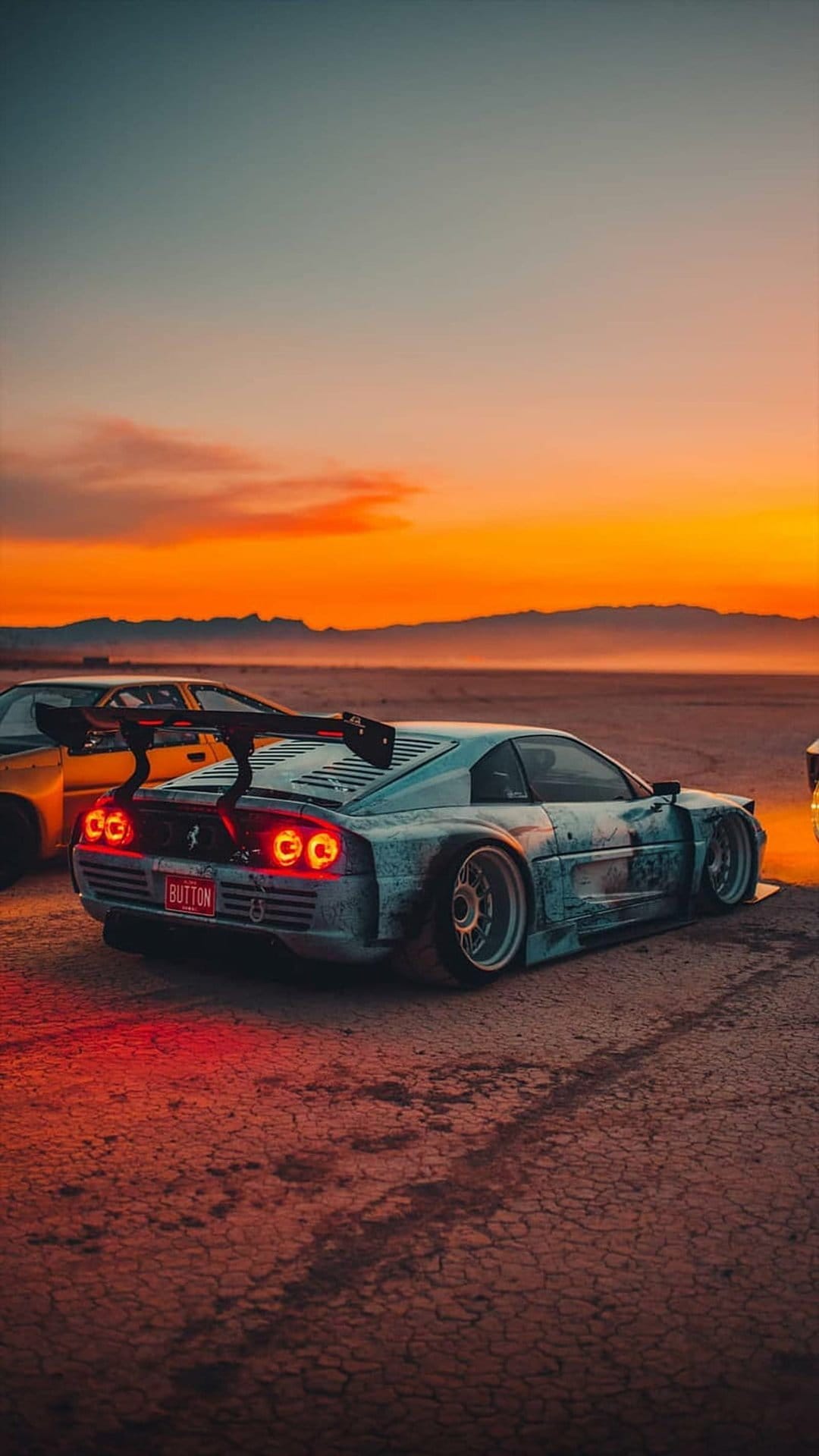 Blue Ferrari with a widebody kit and glowing red taillights parked on a cracked desert floor at sunset.