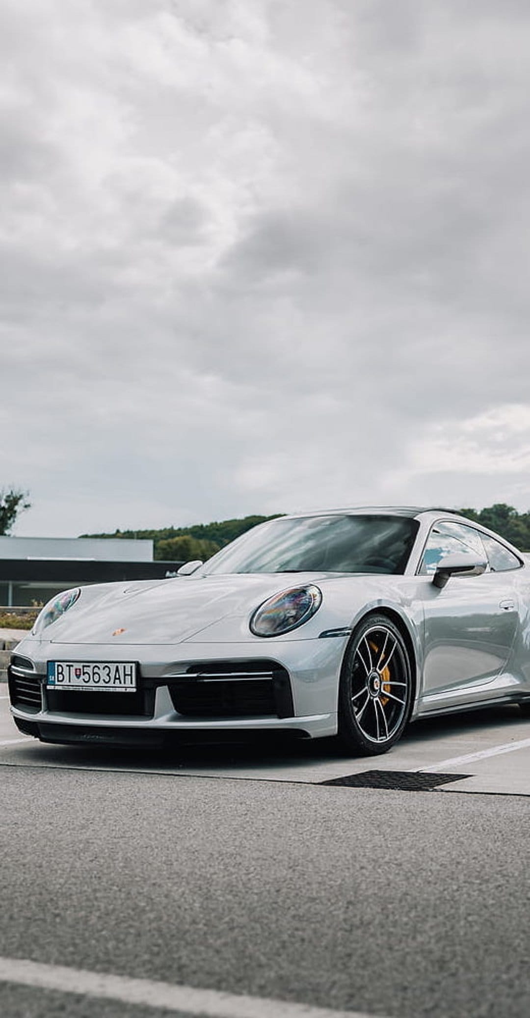 Silver Porsche 911 Turbo S with yellow brake calipers parked on pavement under a moody cloudy sky.