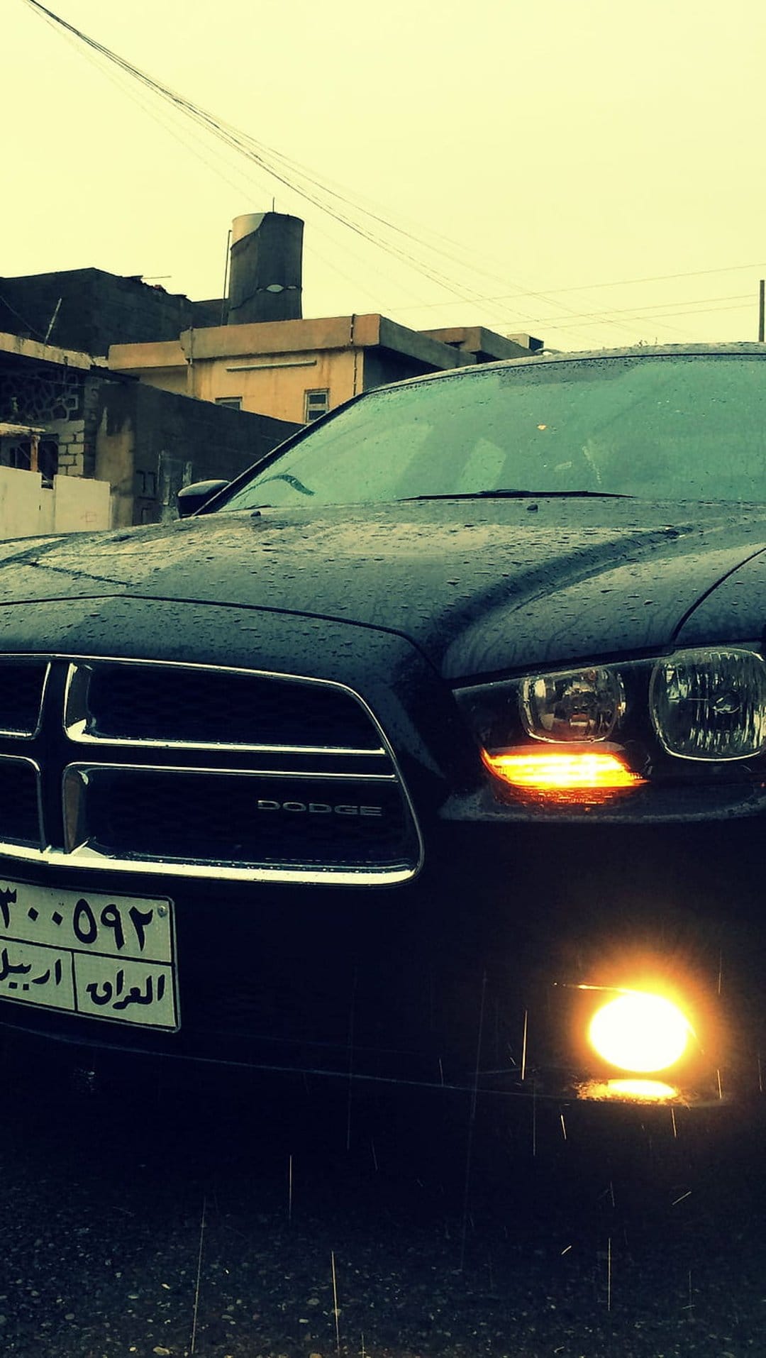 Dodge Charger with glowing headlights on a wet Iraqi street at night under a cloudy sky and urban silhouettes.