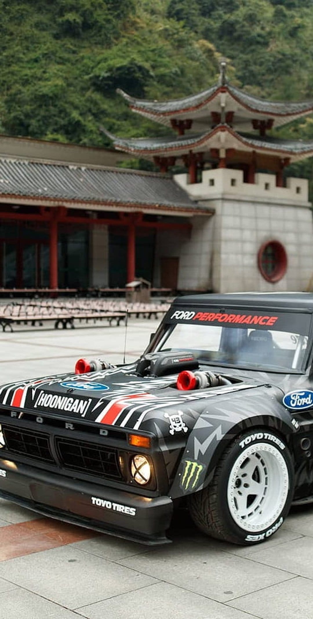 Modified Ford truck with twin turbos parked near a red Asian pagoda with a lush green mountain in the background.