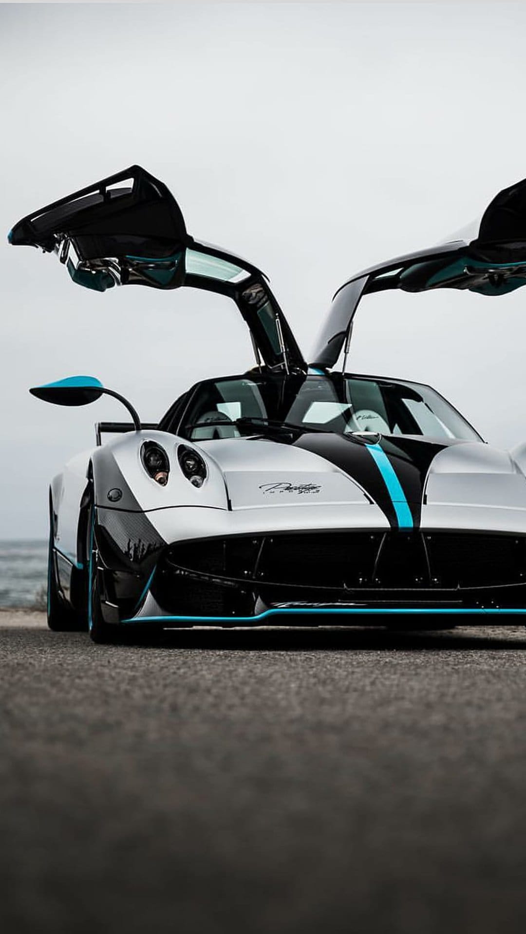 Silver Pagani Huayra Roadster BC with open dihedral doors and blue accents parked near a blurred ocean horizon.