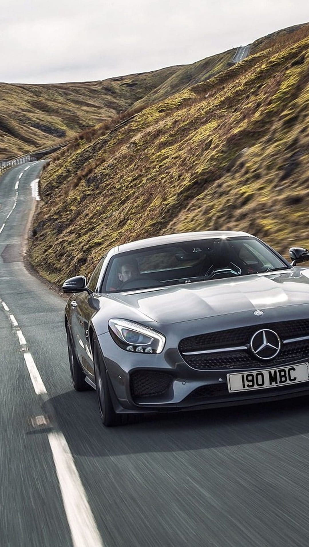 Mercedes-AMG GT S drives on a winding mountain road under an overcast sky surrounded by rolling hills.