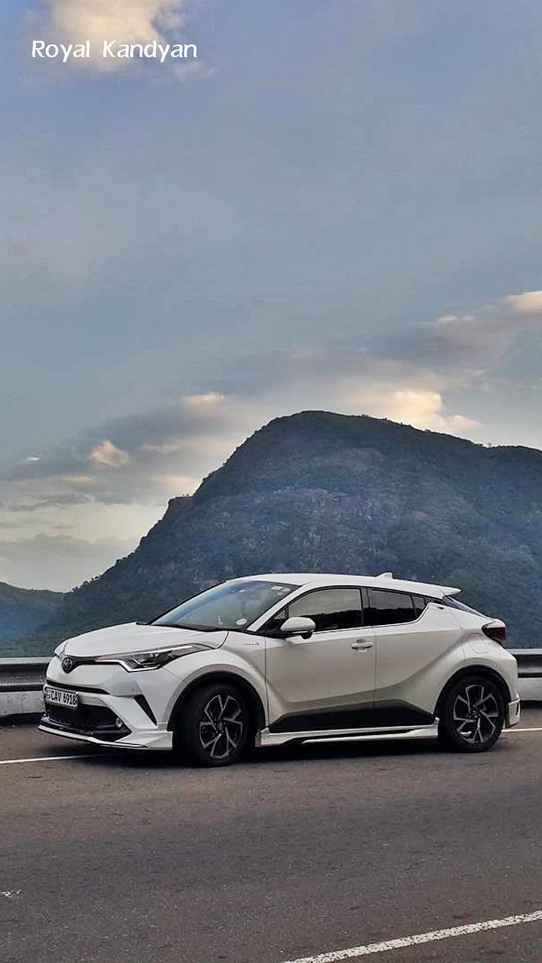 White Toyota C-HR parked on a winding asphalt road with jagged gray mountains and cloudy sky in the background.
