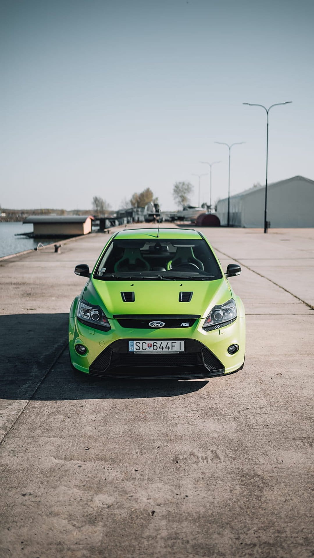 Lime green Ford Focus RS parked on a concrete pier next to calm water with industrial buildings and a blue sky.