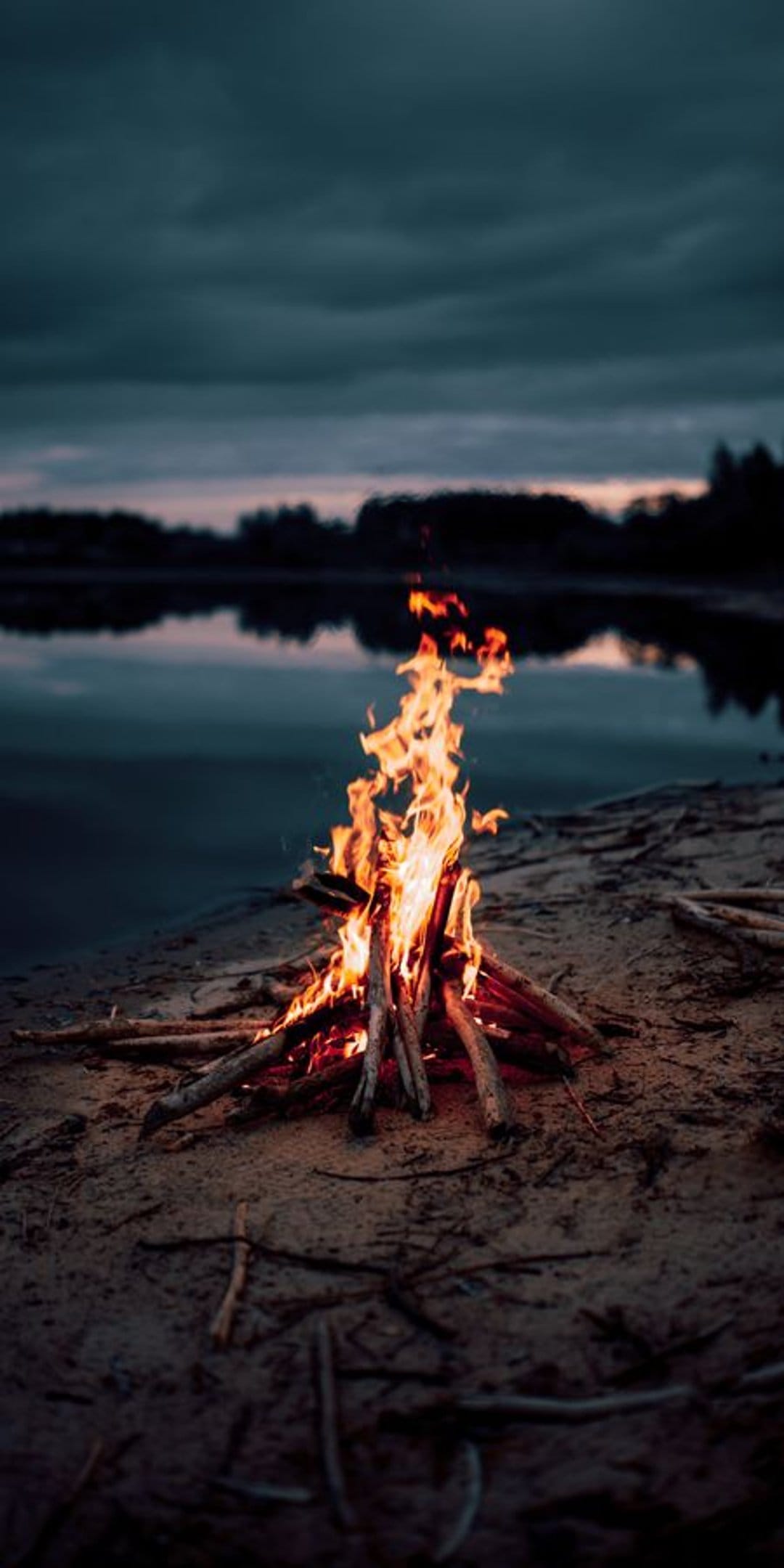 Orange campfire flames flicker on a sandy river shore against a dusky blue sky and calm water at twilight.