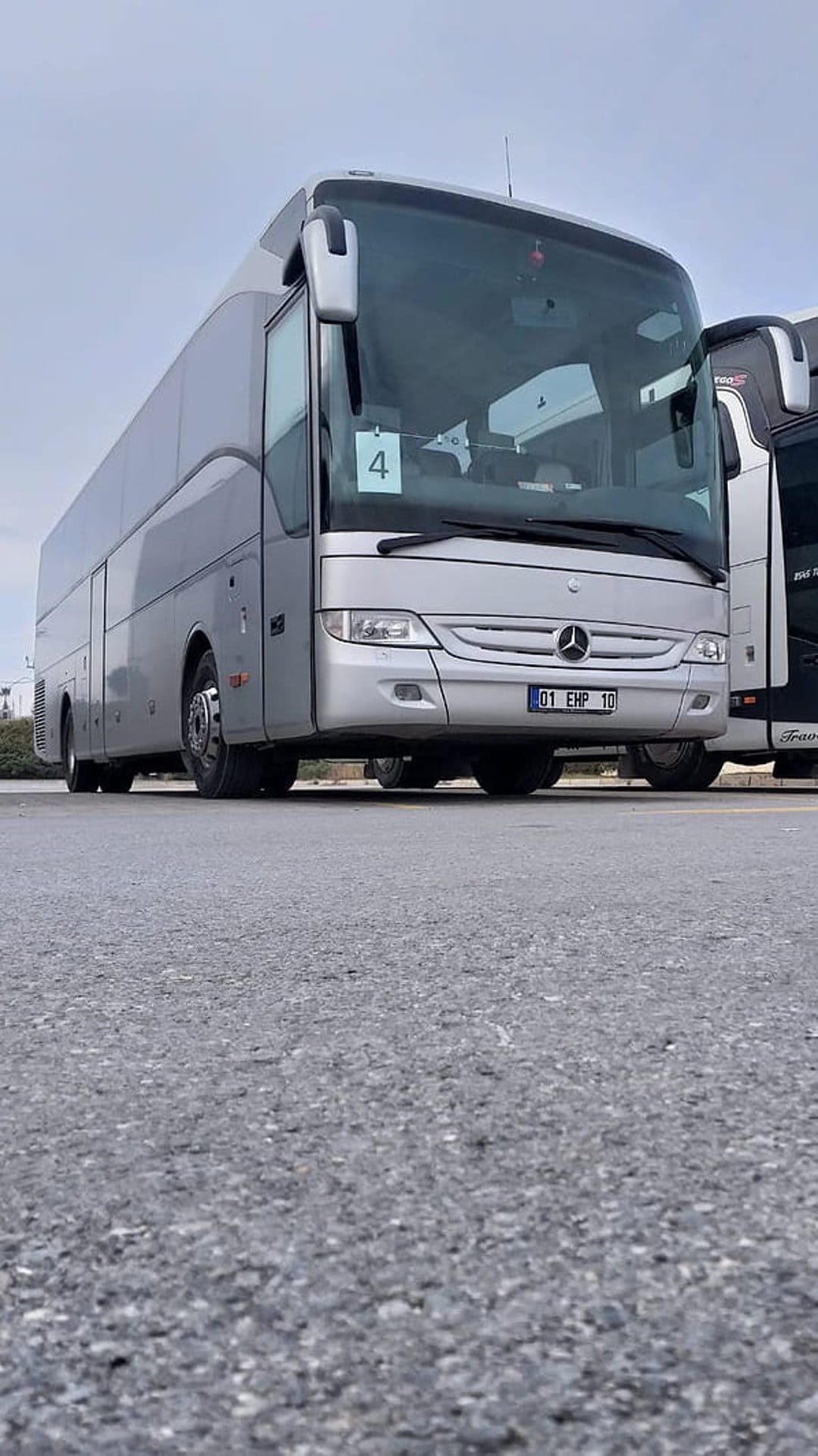 Mercedes-Benz tour bus in silver metallic paint parked on asphalt under a cloudy sky with visible grille logo.