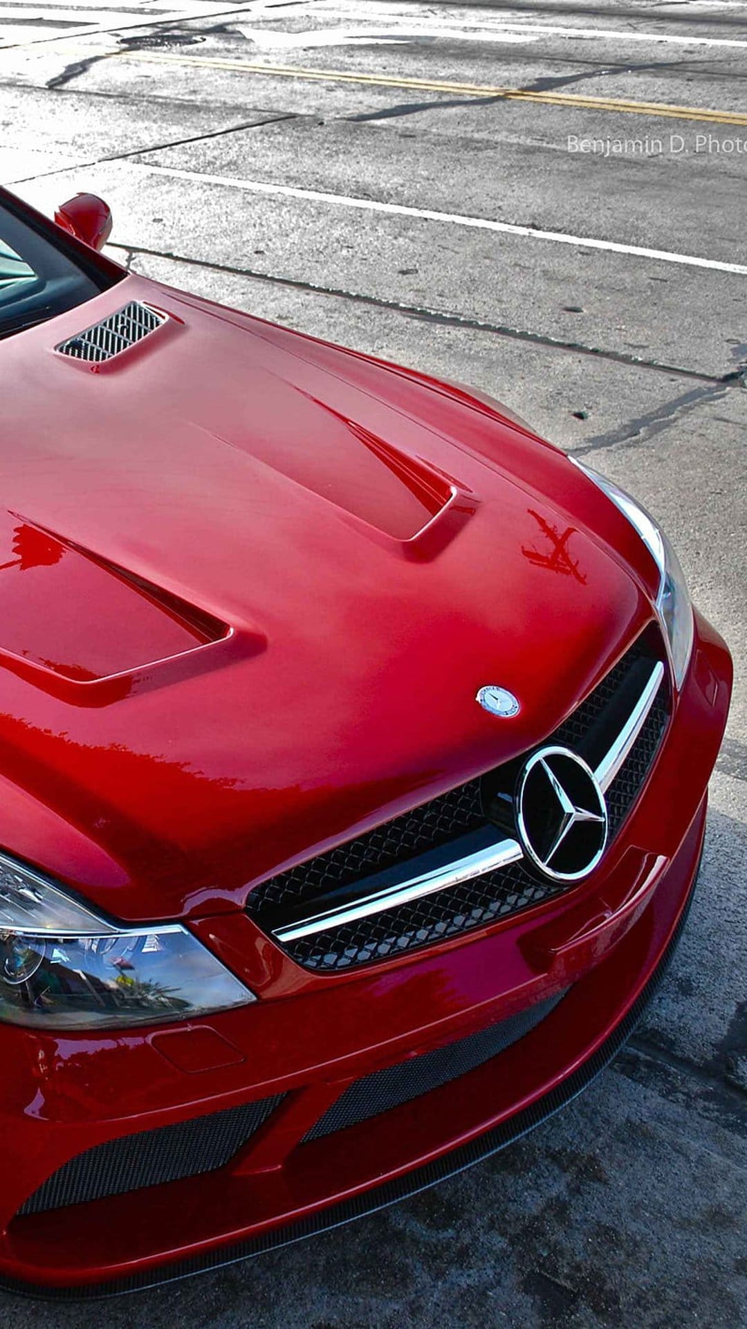Red Mercedes-Benz SL-Class front grille and silver emblem on a sunlit asphalt street with vertical framing.