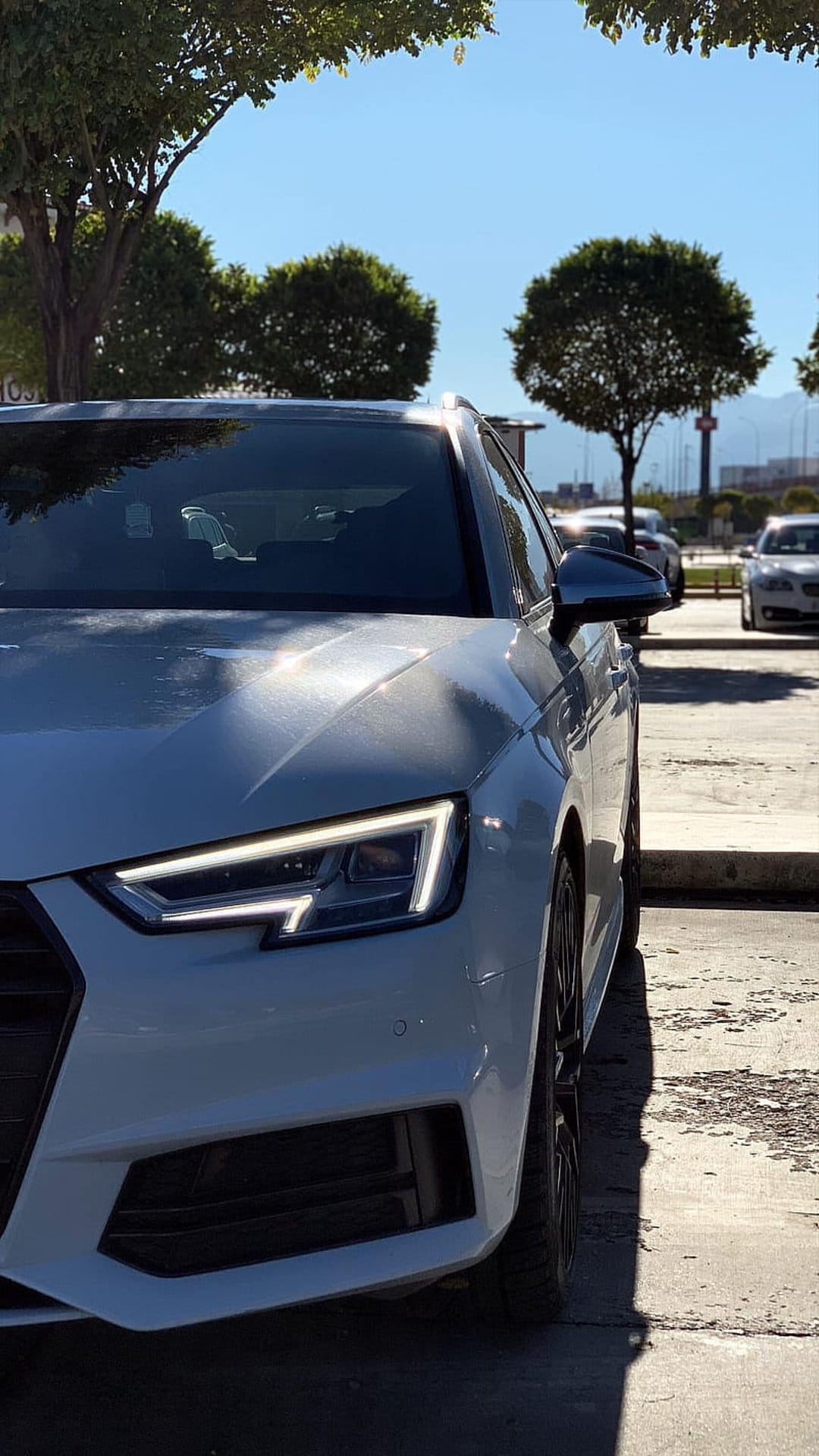 White Audi wagon with glowing LED headlights parked in a sunny lot under a blue sky and green trees.