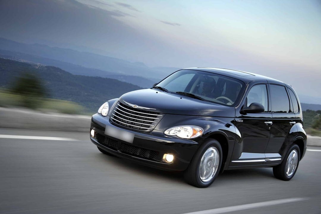 Black Chrysler PT Cruiser driving on a scenic mountain highway with motion blur and a hazy blue sky background.