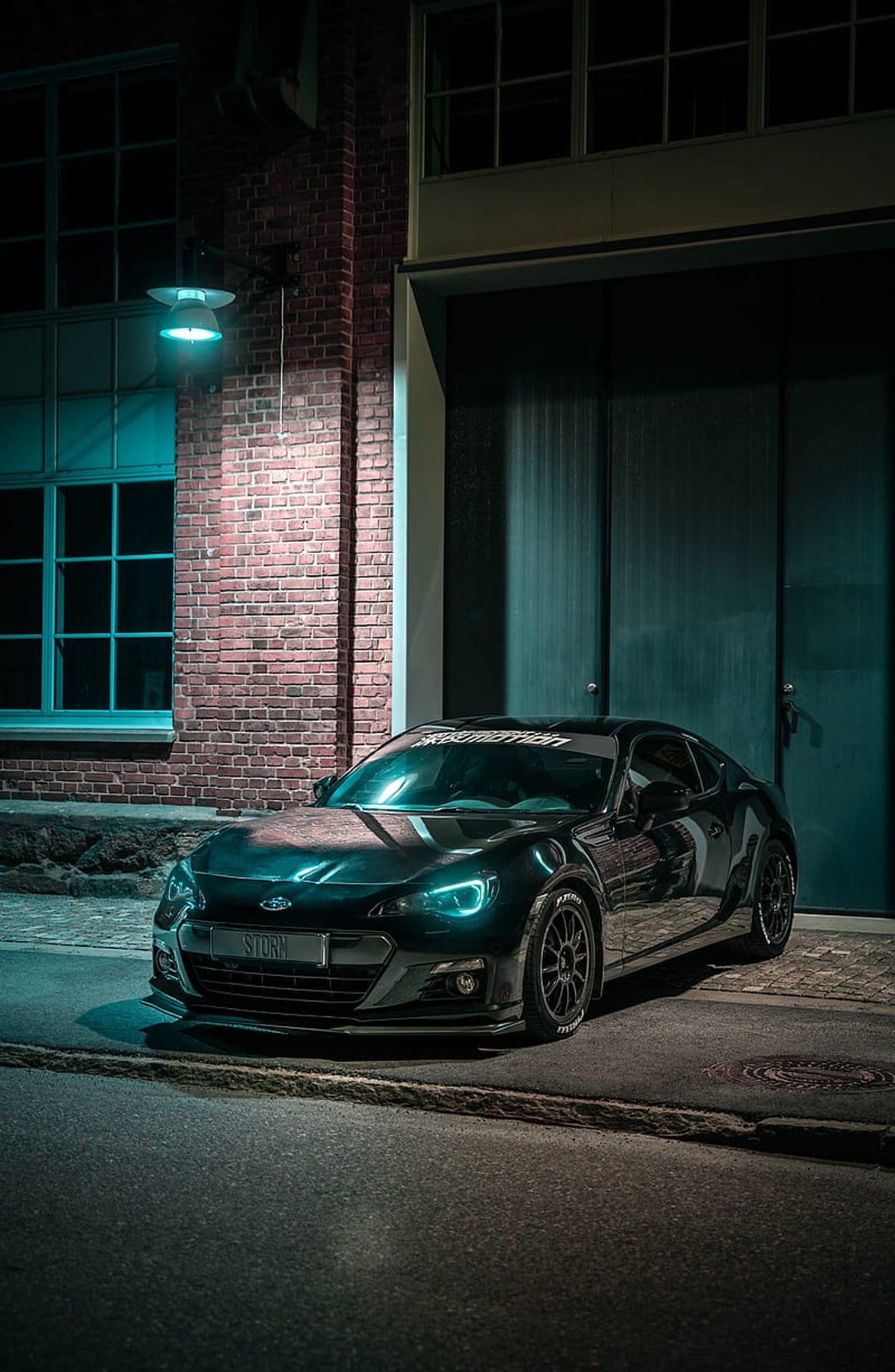 Subaru BRZ with teal headlights parked on a city street at night in front of an industrial brick building.