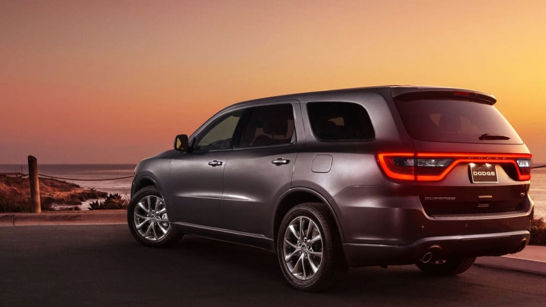 Gray Dodge Durango SUV with glowing red taillights parked on a coastal road overlooking a sunset ocean.