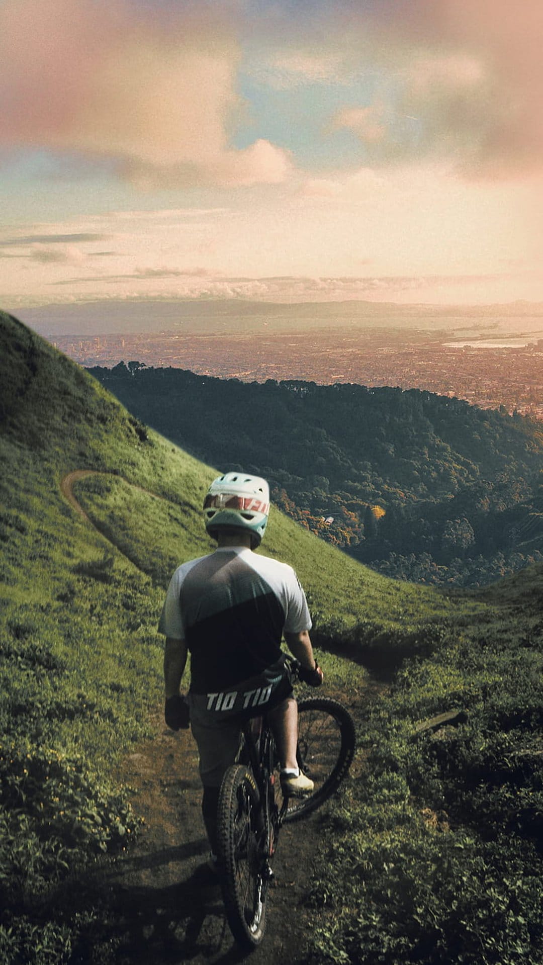 Mountain biker in a helmet stands on a grassy trail overlooking a vast city during a soft golden hour sunset.