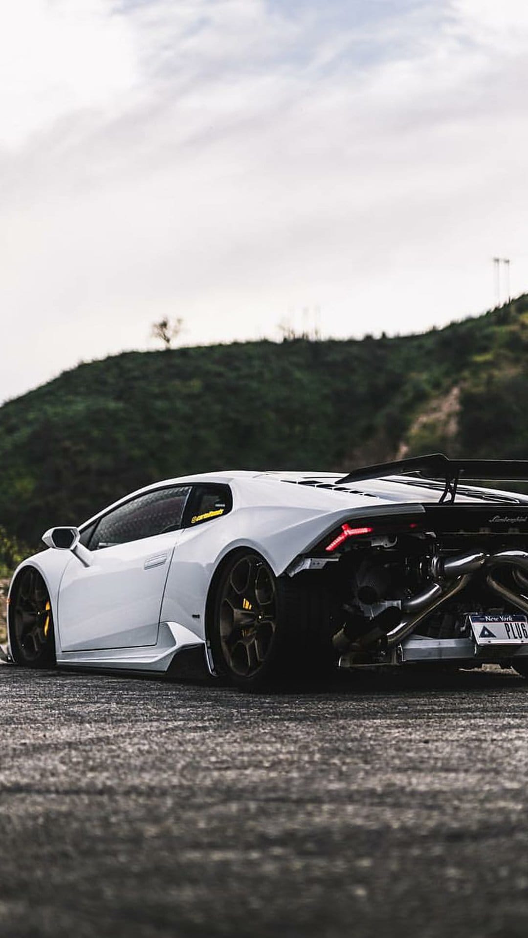 White Lamborghini Huracan with an exposed engine and custom exhaust parked on a road near rolling green hills.