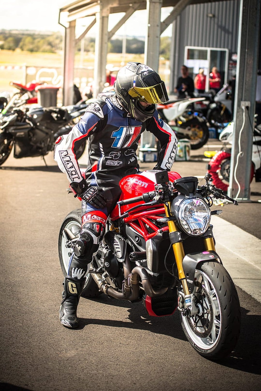 Ducati motorcycle rider in red and blue racing leathers sits on a sportbike in a sunlit pit lane area.