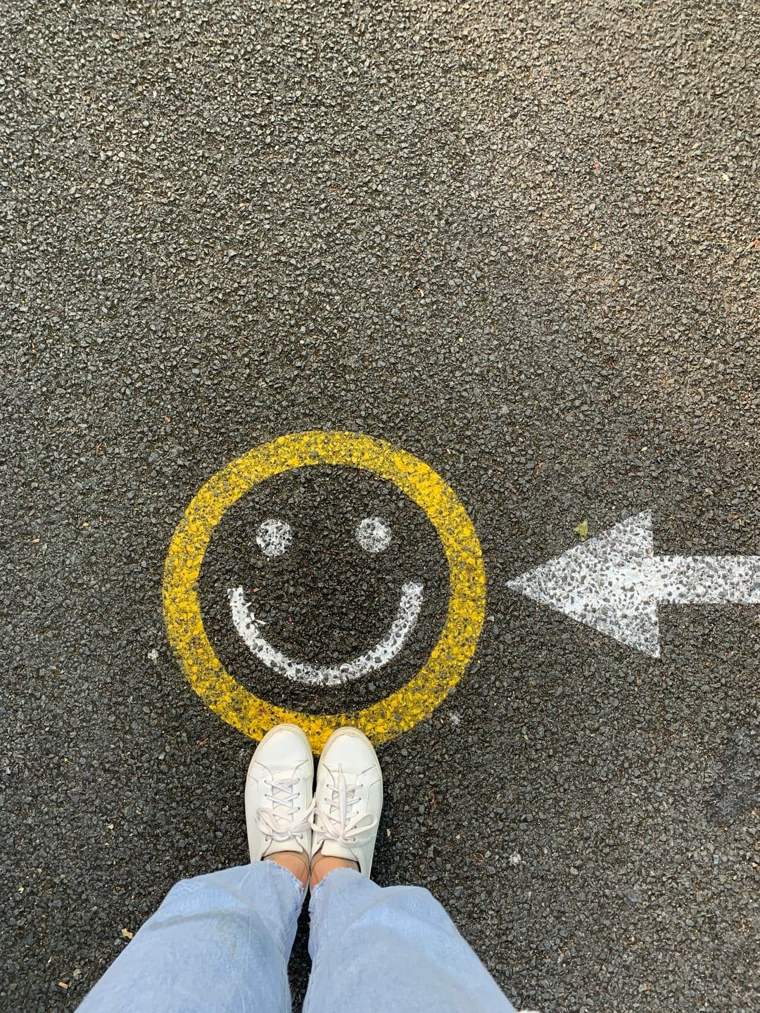 Yellow smiley face and white arrow painted on asphalt with white sneakers standing at the bottom edge.