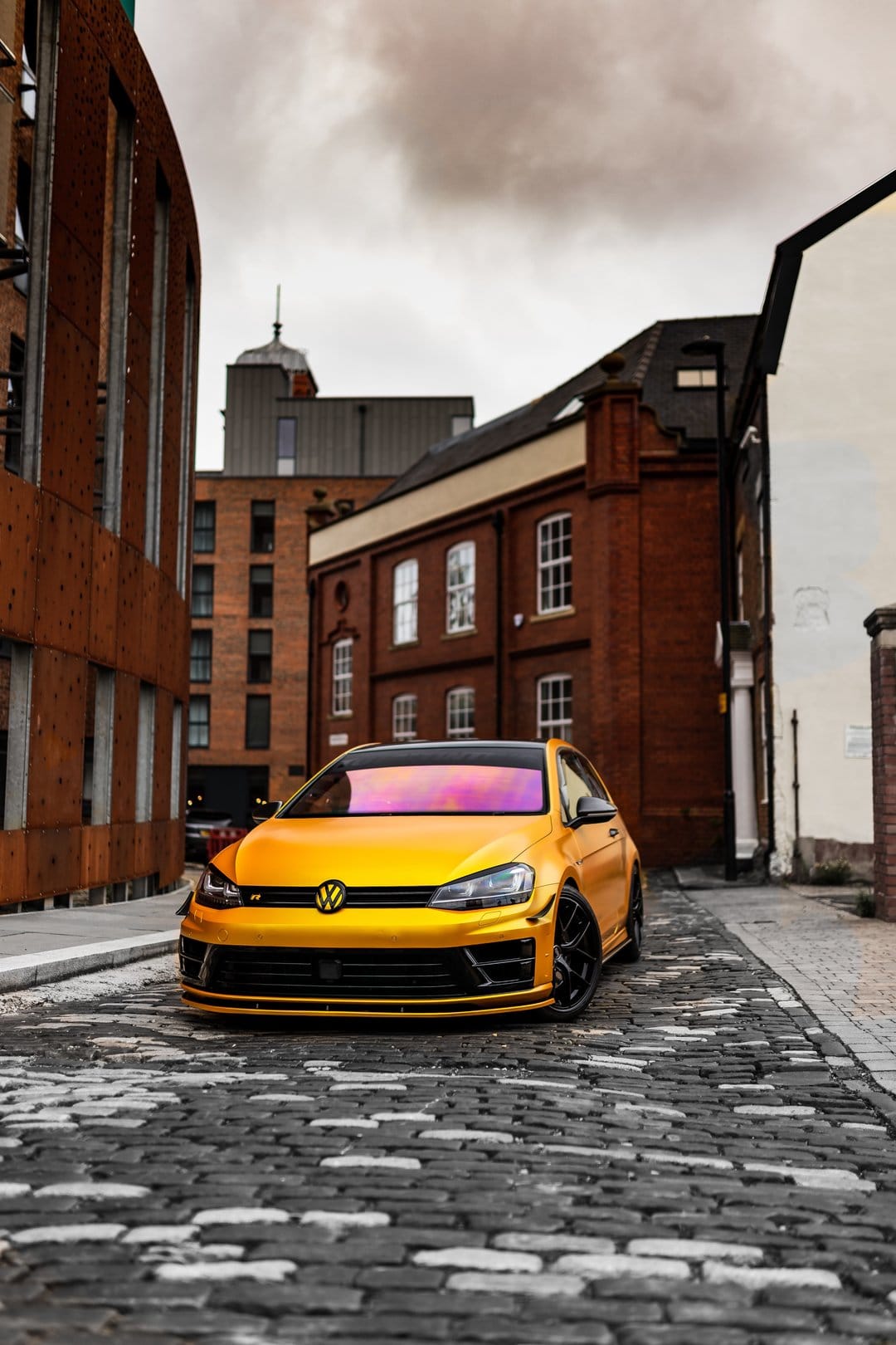 Golden Volkswagen Golf R parked on a grey cobblestone street between industrial brick buildings under cloudy skies.