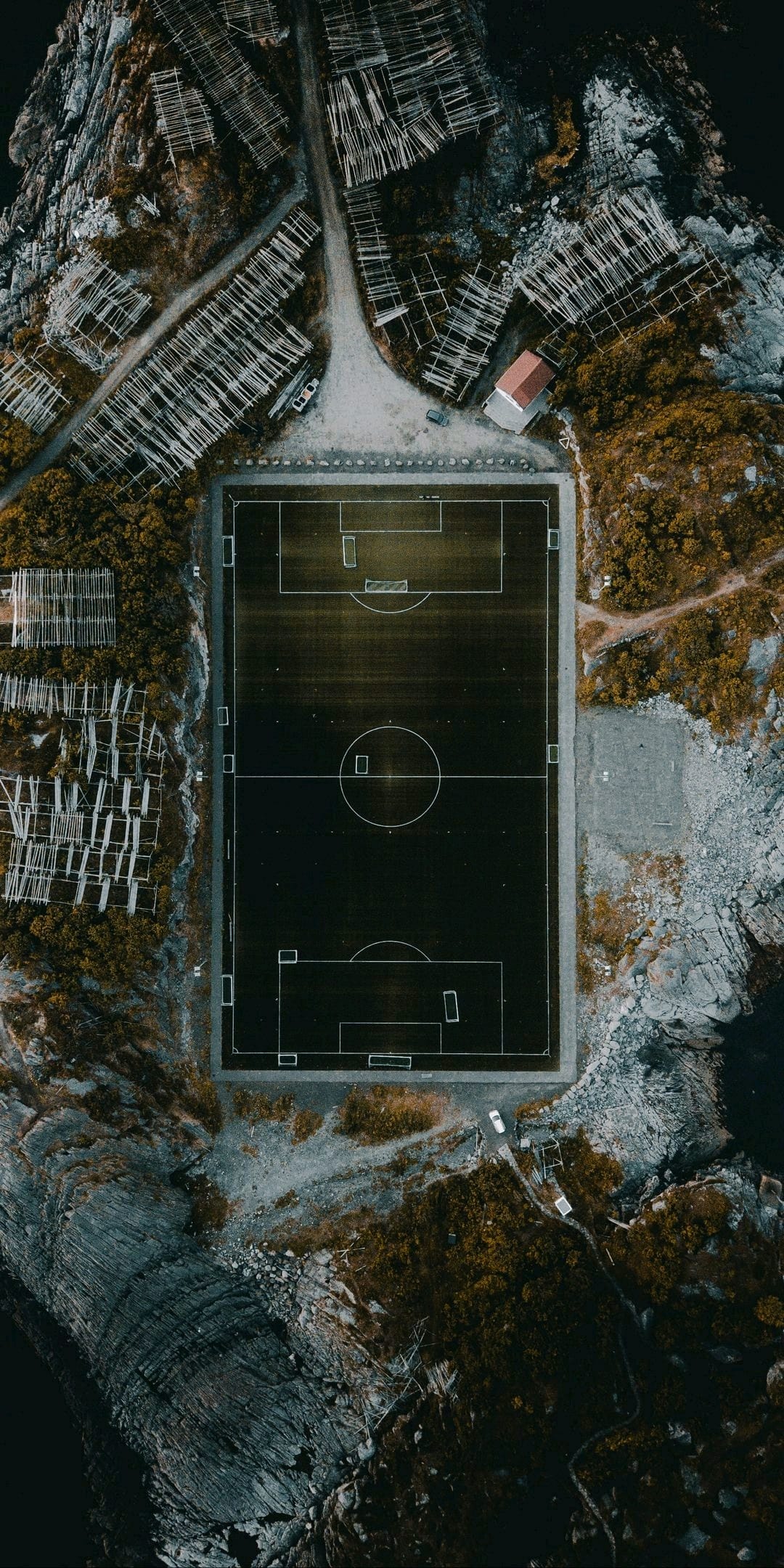 Green soccer field surrounded by rocky terrain and wooden fish racks on a jagged coastline beside the ocean.