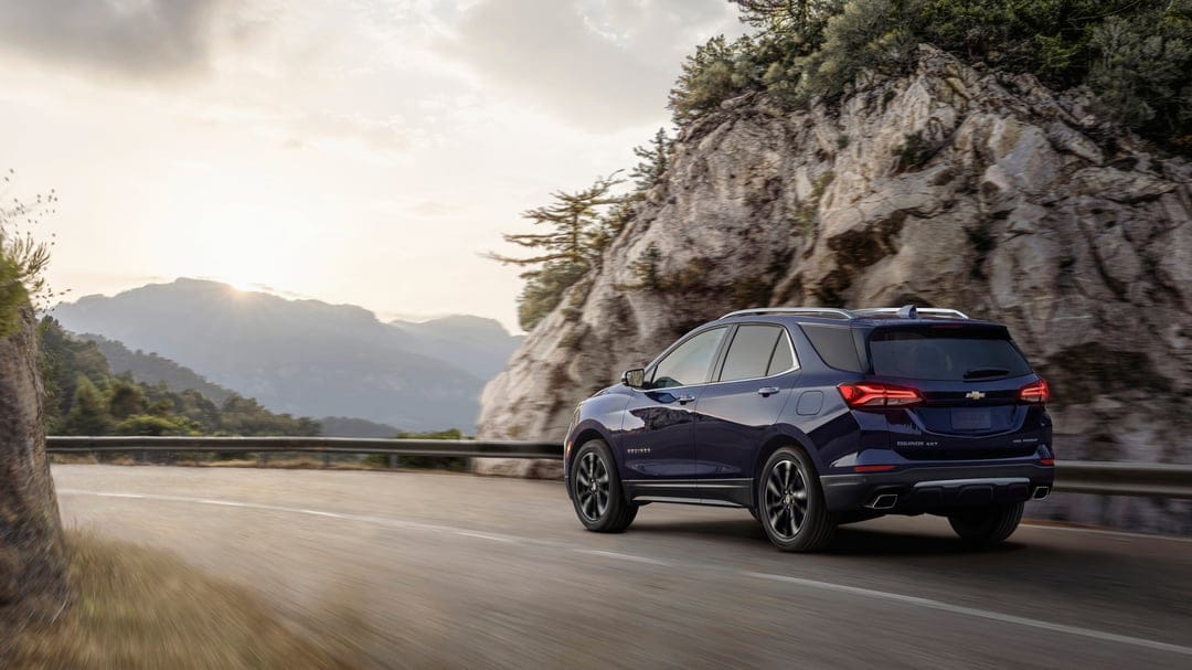 A dark blue SUV driving on a winding mountain road next to a rocky cliff during a hazy golden sunset.