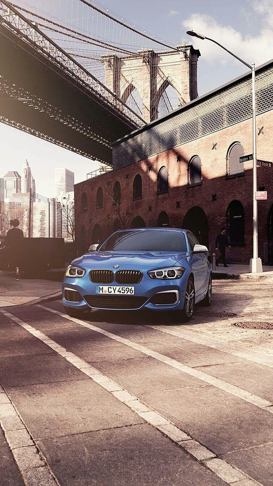 Blue BMW sedan parked on a cobblestone street with the Brooklyn Bridge and brick buildings in the background.