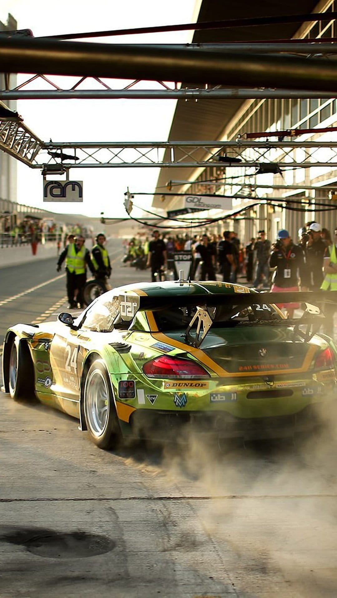 BMW race car with green and yellow livery exits the pits amidst a thick cloud of white smoke on a sunny track.