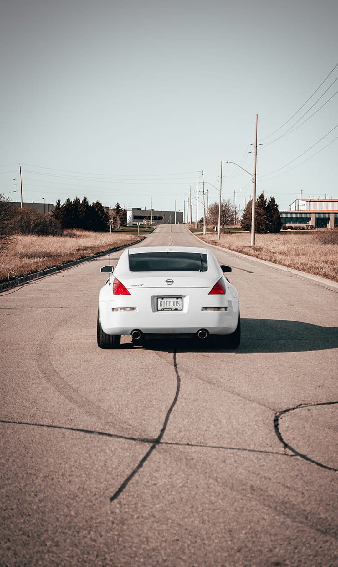 White Nissan 350Z parked on asphalt with black tire marks near dry grass and industrial utility poles.
