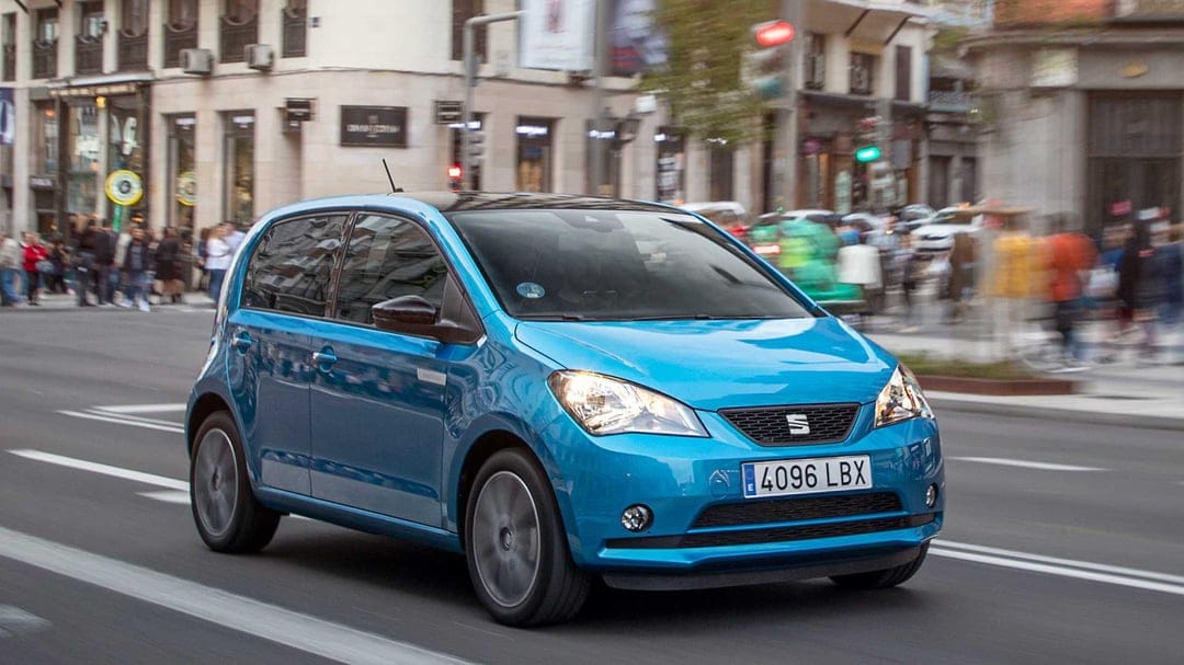 Blue SEAT Mii electric car driving on a city street with blurred buildings and pedestrians in the background.