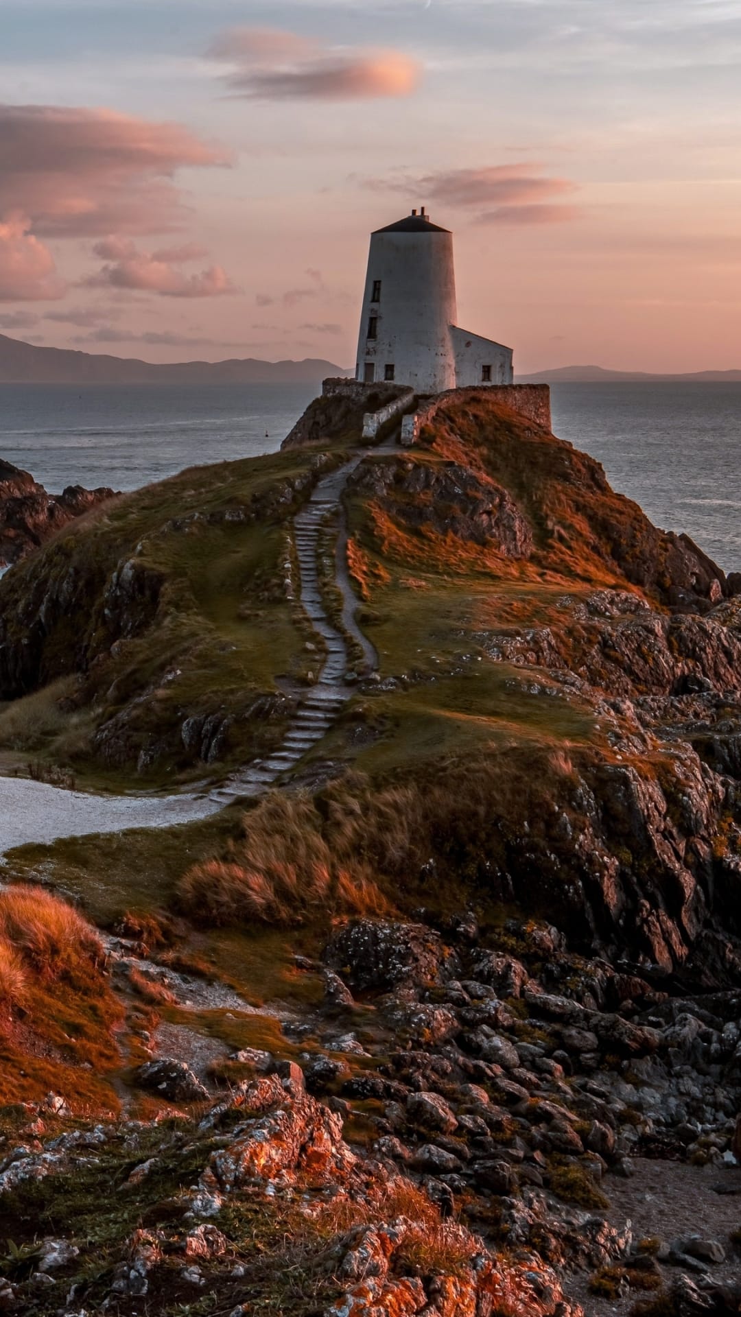 White historic lighthouse and stone steps on a grassy cliff at sunset overlooking a calm sea in Anglesey, Wales.