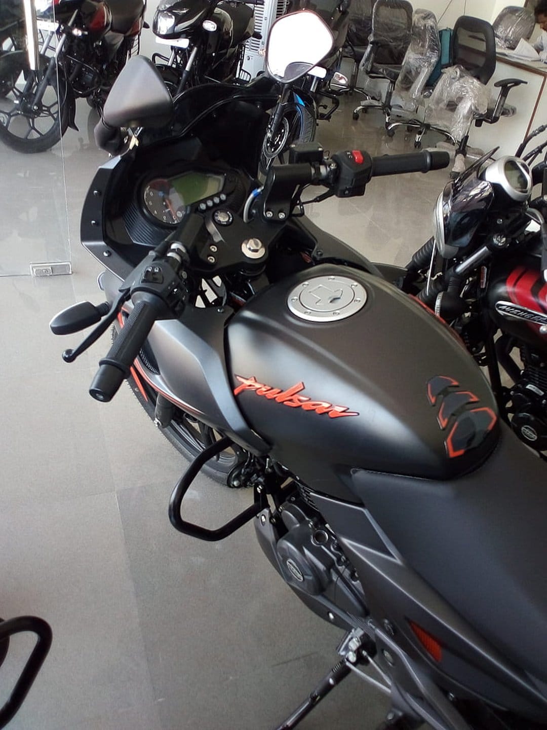 Matte black motorcycle fuel tank with a vibrant orange Pulsar logo, featuring handlebars and a side mirror.