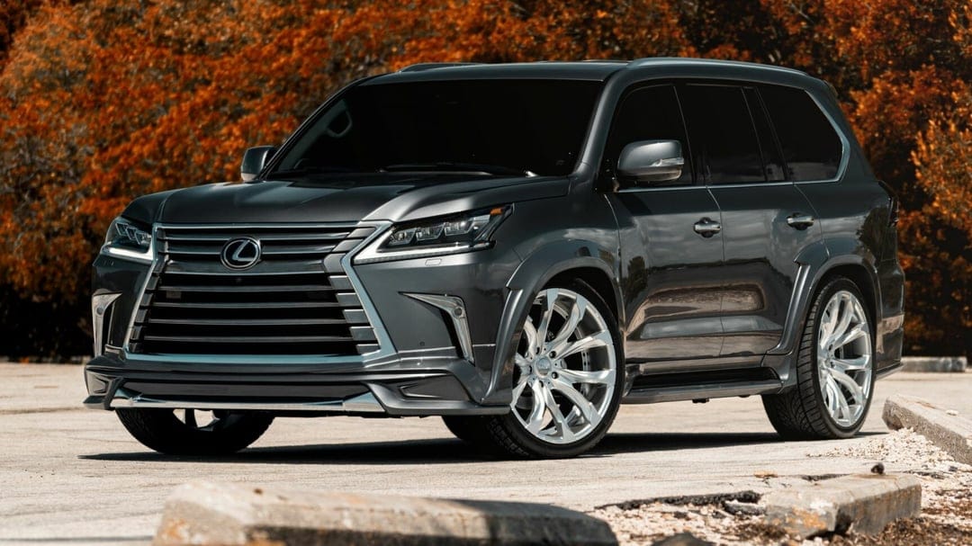 Dark grey Lexus LX SUV with custom silver wheels parked on concrete against a backdrop of orange autumn trees.