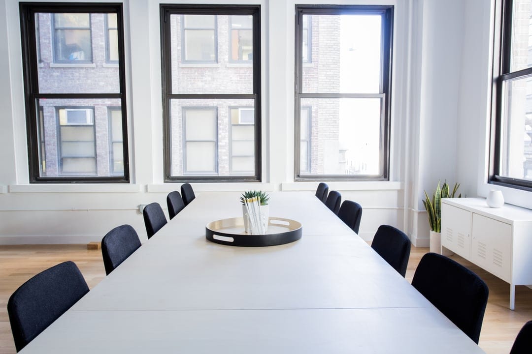 White conference table and black chairs in a sunlit office room with windows overlooking a blurred city skyline.