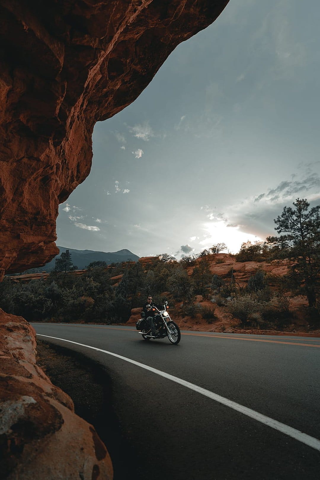 Motorcyclist riding a cruiser on a desert highway between tall red sandstone formations under cloudy skies.