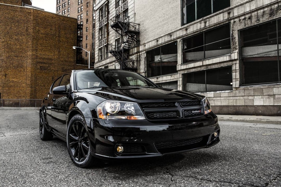 Black Dodge Avenger parked on a gritty urban street with industrial brick buildings and a fire escape.