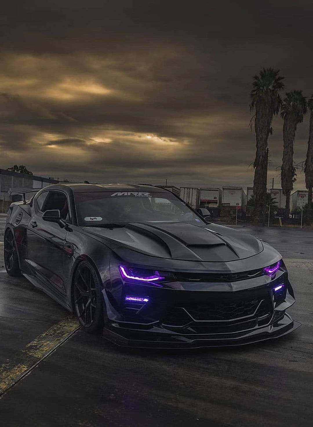 Black Chevrolet Camaro with purple LED headlights parked outdoors at dusk under a cloudy sky with palm trees.