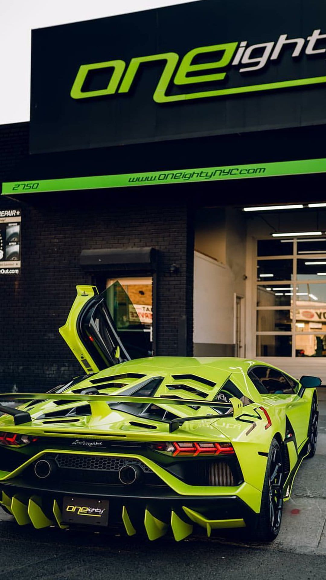 Lime green Lamborghini Aventador SVJ with open scissor doors parked in front of a brick urban garage entrance.