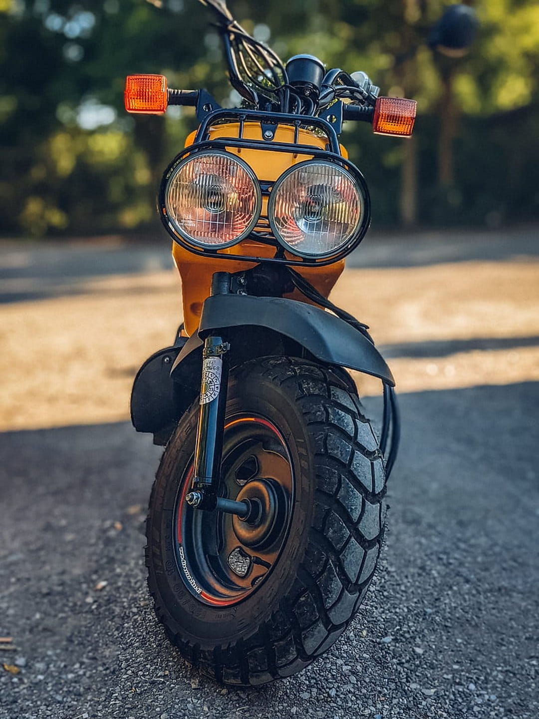 Yellow scooter with dual round headlights and knobby tires sits on gravel against a blurred green forest backdrop.