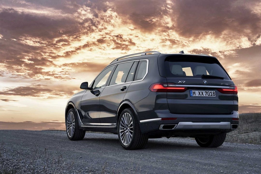Dark BMW X7 SUV parked on a gravel path against a dramatic sunset sky with golden clouds and glowing LED taillights.