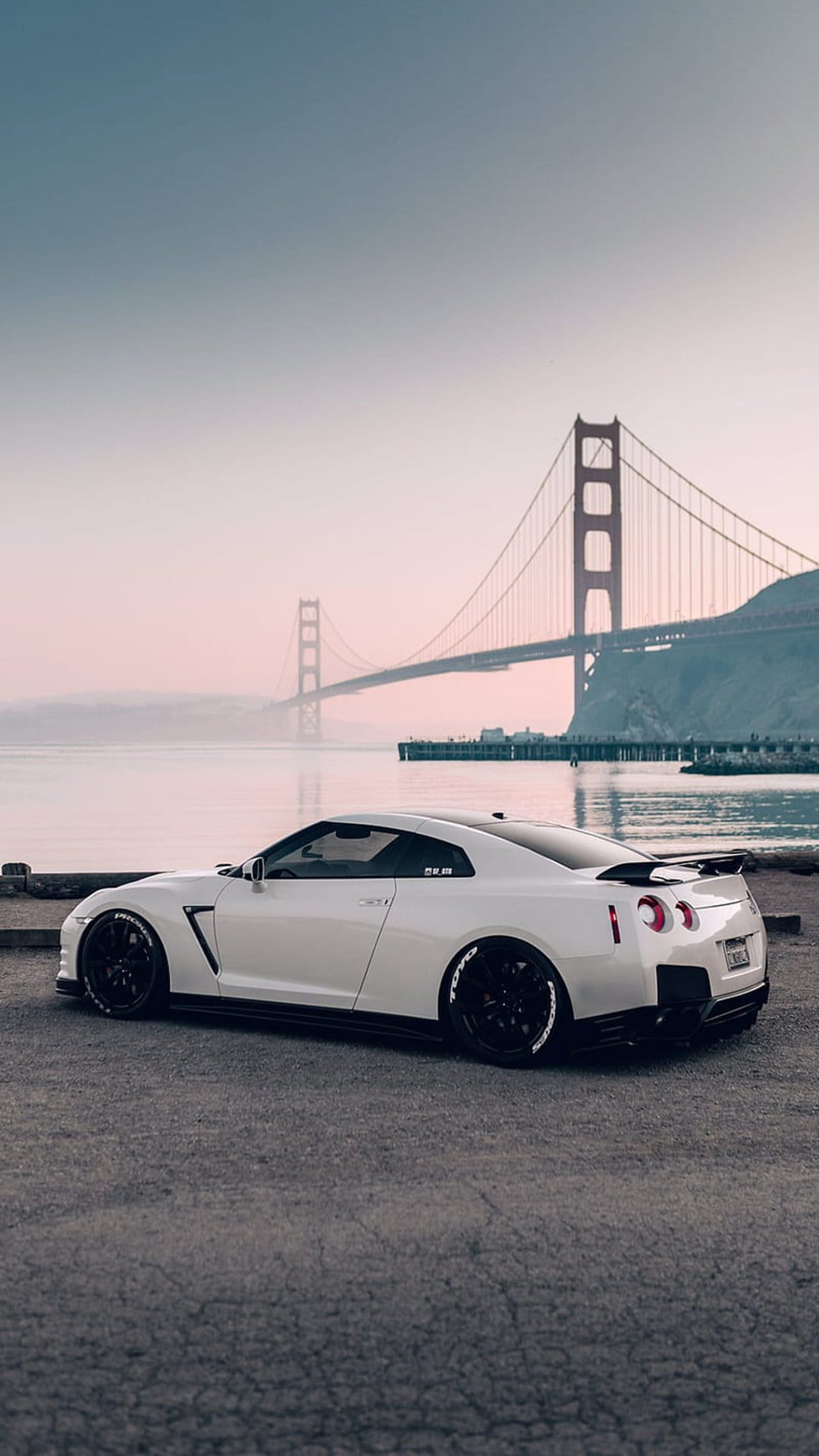 White Nissan GT-R sports car parked on a roadside with the red Golden Gate Bridge visible through morning mist.