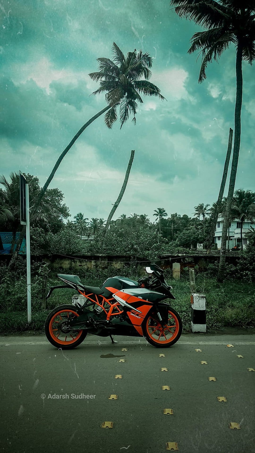 Orange KTM sportbike parked on a wet road with leaning palm trees and a dark cloudy sky in the background.