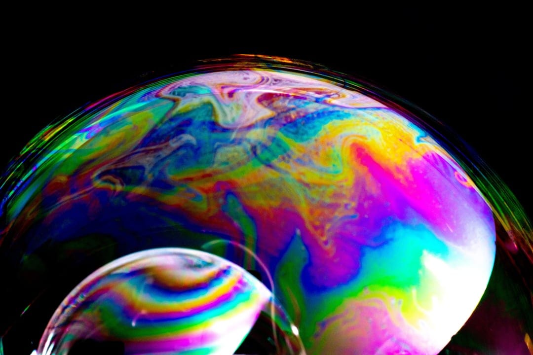 Iridescent soap bubble film displays swirling rainbow patterns and liquid textures against a dark background.