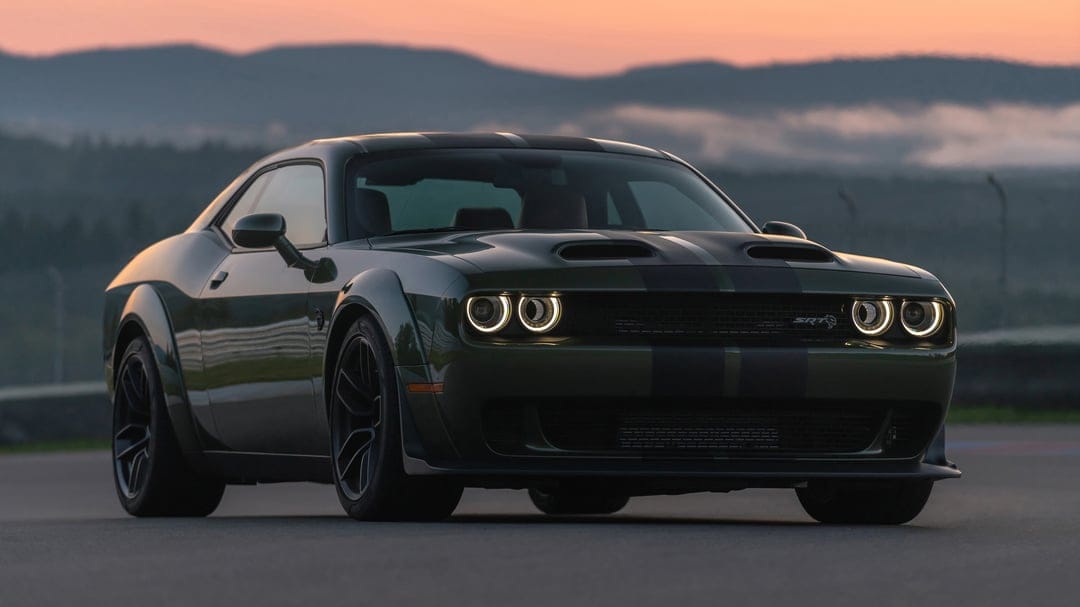 Dark green Dodge Challenger SRT Widebody with glowing halo headlights on a track at sunset with misty mountains.