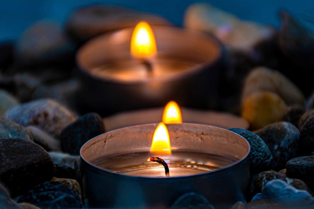 Two lit tea light candles glow amongst smooth dark river stones with a soft bokeh background for tranquility.