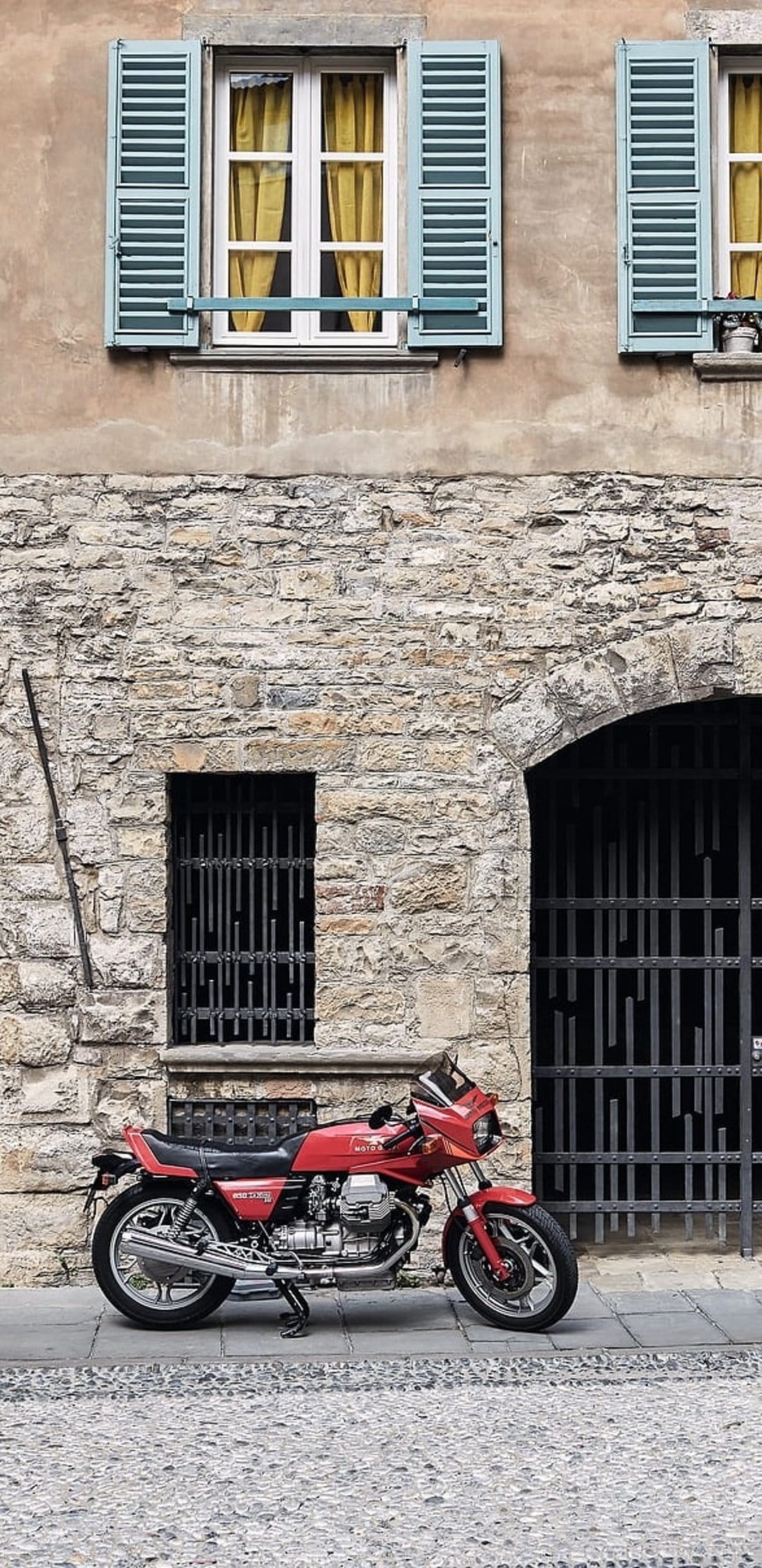 Vintage red motorcycle parked on a cobblestone street before a stone building with light blue window shutters.